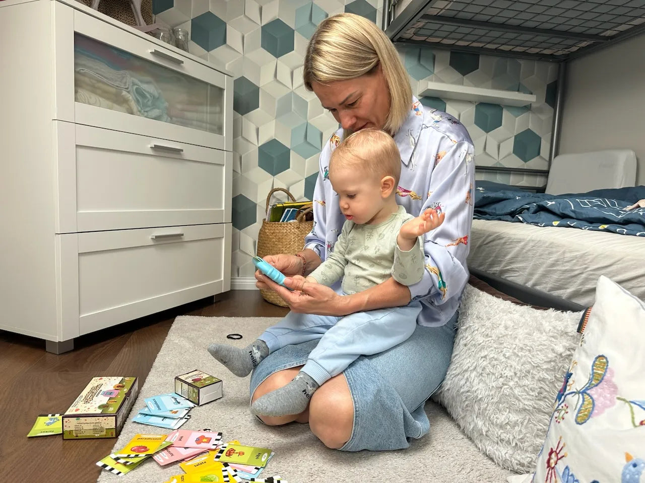 Mamička učí dieťa nové slová pomocou Kinderspeech čítačky v detskej izbe s rozloženými kartičkami na koberci.